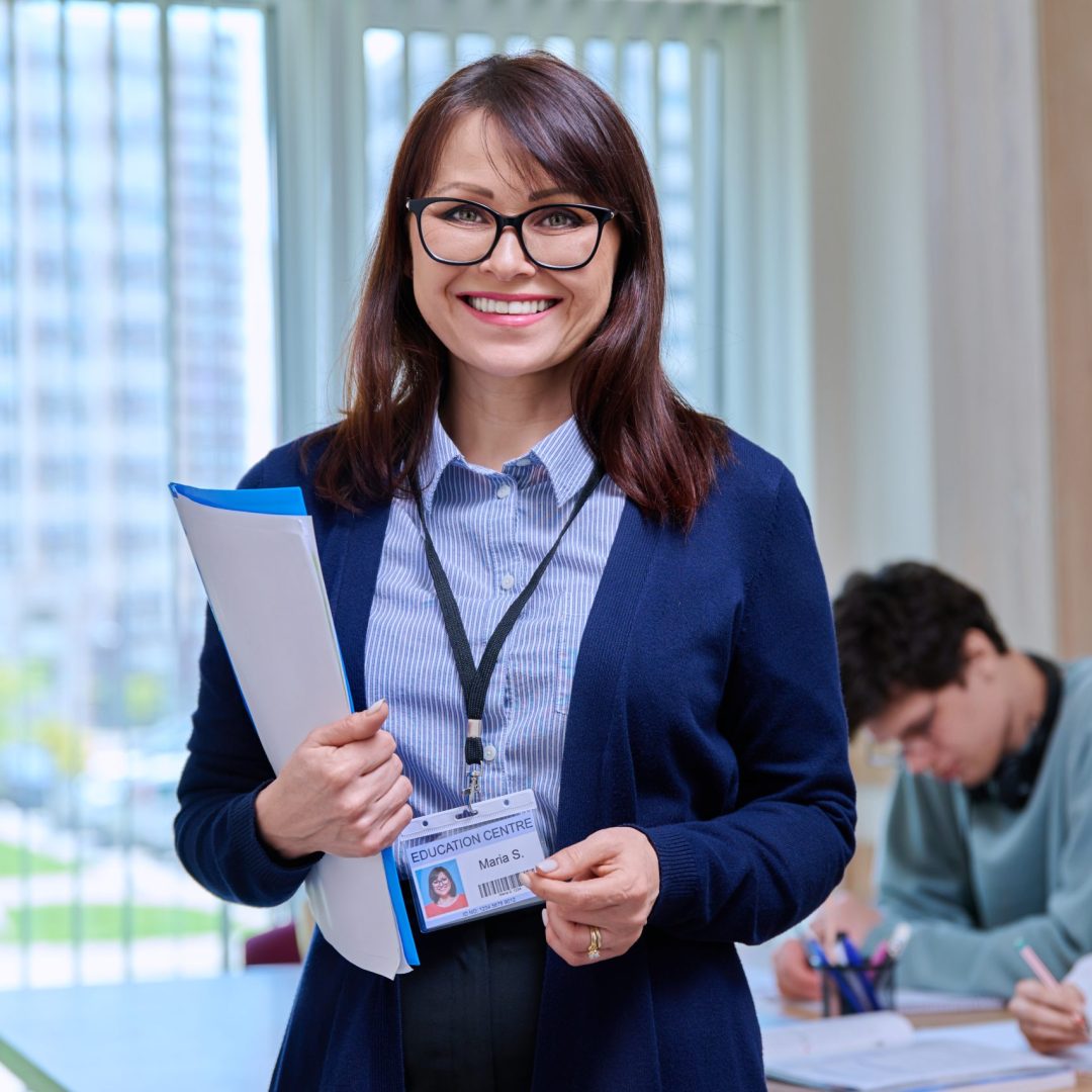 Portrait of confident middle aged female teacher schoolmaster tutor educator looking at camera, in college classroom with students at desk. Education, learning, language courses, knowledge concept
